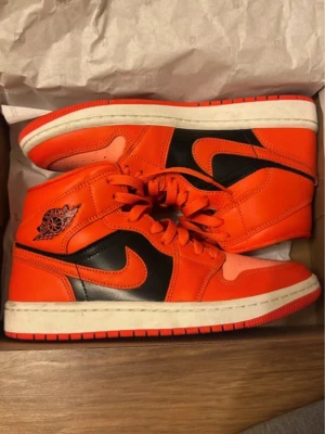 Orangea Air Jordan 1 Mid sneakers - Säljer ett par Air Jordan 1 Mid sneakers i orange och svart med klassisk Nike swoosh på sidan. Skorna har snörning, hög siluett och är tillverkade i skinn med vita sulor och röda detaljer. Perfekta för dig som vill sticka ut med din stil. Använda 1 gång. INTE creasade!