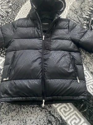 Svart pufferjacka från Alessandro - Snygg svart pufferjacka från Alessandro med diskret mönster i tyget och stora dragkedjor. Jackan har huva, två sidofickor med dragkedja och hög krage. Perfekt för kalla dagar och ger en riktigt modern look.