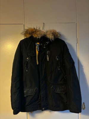 Svart parkasjacka med pälskrage - Snygg svart parkasjacka från Parajumpers med avtagbar huva och fluffig pälskant i beige och brunt. Jackan har flera fickor med dragkedja och knappar, samt den klassiska gula remmen vid halsen. Perfekt för kalla dagar och riktigt varm.