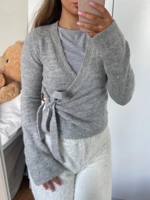 Kofta - Mysig grå kortare omlottkofta med långa ärmar och knytband i sidan. Koftan har ribbade muddar och en mjuk, stickad känsla som passar perfekt till lager på lager. Enkel att matcha med olika outfits för en avslappnad och trendig look. Storlek XS från Gina Tricot 🩶