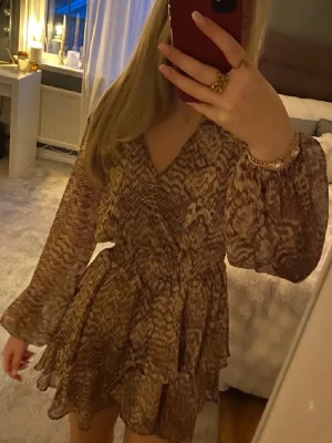 Brun kort klänning med ormmönster Zara - Kort klänning från Zara i brun och beige ton med ormmönster. Klänningen har v-ringning, långa puffärmar och volangdetaljer i kjolen. Tillverkad i ett lätt, luftigt material som ger en flowy känsla. Perfekt för dig som gillar trendiga statement-plagg.