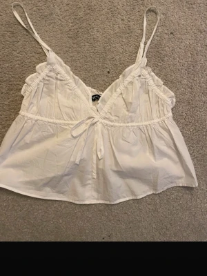 Vit linnetopp med knyt från Gina Tricot - Söt vit linnetopp från Gina Tricot med tunna axelband, v-ringning och volangdetaljer. Toppen har en knytning framtill och är lätt utsvängd nedtill. Perfekt för varma dagar och enkel att styla med jeans eller kjol.