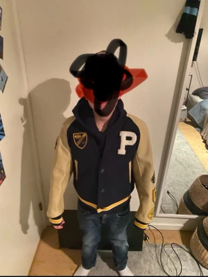 Mörkblå och beige bomberjacka PLC - Snygg mörkblå bomberjacka med beige ärmar och detaljer. Stor P på bröstet och broderad PLC-text på ryggen. Jackan har tryckknappar, ribbade muddar och coola patchar på ärmarna. Perfekt för dig som vill sticka ut med en klassisk college-look.