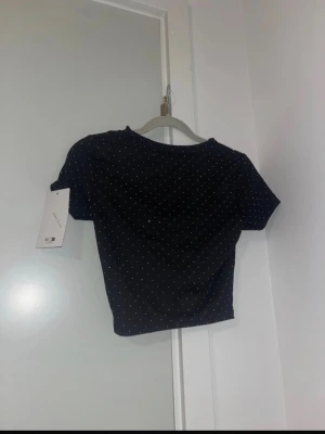 Svart set - Trendig svart topp med kjol som lyser och en snygg ringdetalj framtill. Toppen har korta ärmar och är croppad, vilket ger en modern look. Perfekt för dig som gillar att sticka ut med detaljer. Materialet känns mjukt och följsamt. Kjolen har en volang med en guld detalj.