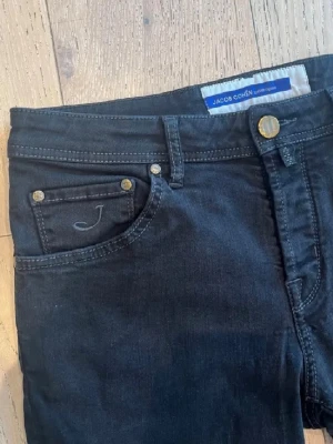 Jacob Cohën Scott jeans strl W31 - Snygga svarta jeans från Jacob Cohën med klassisk femficksmodell och diskreta sömmar. Modellen Scott, vilket är en perfekt slim fit. Patchen i mocka har tyvärr gått sönder lite under tvätten. Tillverkade i mjukt denimtyg med stretch för extra komfort. Modellen är 182 cm 70 kg. Hör av er vid frågor och funderingar!