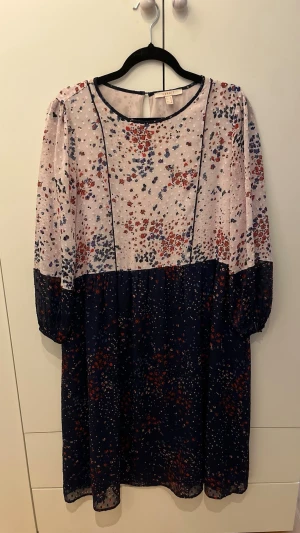 Blommig långklänning - Söt långklänning med blommigt mönster i blått, vitt och rött. Klänningen har lång ärm med resår vid ärmslut, rund halsringning och lätt transparent material. Perfekt för dig som gillar romantisk och avslappnad stil.