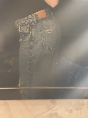 Blå slitna mom jeans med hål - Säljer ett par blå ”boyfriend” jeans med slitningar och hål på knäna. Jeansen har hög midja, raka ben och klassiska fem fickor. Materialet är denim i bomull och de har en avslappnad vintage-look. Perfekta för en trendig och avslappnad stil.