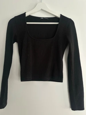 Svart ribbad topp från Zara - Snygg svart ribbad topp från Zara med lång ärm och fyrkantig urringning. Toppen är croppad och har en tight passform som framhäver figuren. Perfekt att styla med höga jeans eller kjol för en trendig look.