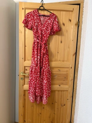 Röd blommig långklänning med knyt - Snygg röd långklänning med vita blommor och v-ringning. Klänningen har korta ärmar, ett matchande knytband i midjan och ett luftigt fall. Perfekt för sommarens alla tillfällen och ger en romantisk vibe.