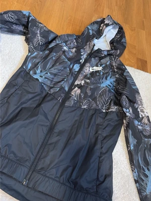 Nike vindjacka med bladmönster - Snygg vindjacka från Nike med svart bas och bladmönster i grått och blått upptill. Jackan har hel dragkedja, huva och meshfoder för extra komfort. Nike-logga i vitt på bröstet. Perfekt för dig som vill ha en sportig och trendig look.