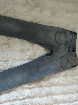 Grå tvättade jeans från Jack&jones - Säljer ett par grå tvättade jeans från Jack&jones med raka ben och klassisk femficksmodell. Jeansen har snygga slitningar och en avslappnad passform. Materialet är bomull med en mjuk känsla och de har en patch med logga bak i midjan.