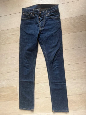 Mörkblå jeans från Nudie Jeans - Snygga mörkblå jeans från Nudie Jeans med klassisk femficksmodell och orange kontrastsömmar. Jeansen har knappgylf och raka ben. Tillverkade i kraftigt denimtyg som ger en tidlös look. Perfekta för dig som gillar stilrena och enkla jeans.