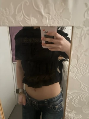 Svart croppad spetsblus - Säljer en svart croppad blus med spetsdetaljer och volanger. Toppen är genombruten och har korta ärmar, perfekt för dig som gillar en edgy och trendig stil. Passar till både jeans och kjol för en snygg look.