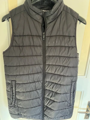Svart dunväst från Everest - Snygg väst från Everest med en liten krage. Västen har dragkedja framtill och två fickor på sidorna. Perfekt för våren och sommaren. Lätt att matcha. Storlek 170-176 (passar som S). 
