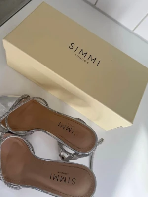Silvriga klackar med strass från Simmi London - Säljer ett par snygga silvriga pumps från Simmi London med tunna remmar dekorerade med strass. Skorna har öppen tå och smal klack, perfekt för dig som gillar en glittrig och trendig look. Materialet är syntet med metallicfinish och strassdetaljer.