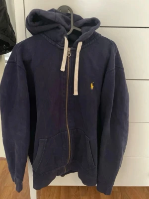 Mörkblå hoodie från Polo Ralph Lauren - Mörkblå hoodie med dragkedja från Polo Ralph Lauren. Klassisk gul broderad logga på bröstet, vita snören i huvan och två stora fickor framtill. Tillverkad i mjuk bomull, perfekt för en avslappnad och stilren look.