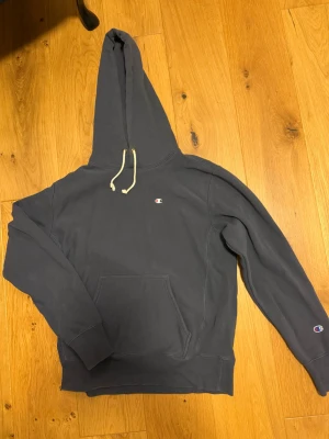 Mörkblå hoodie från Champion - Säljer en mörkblå hoodie från Champion med klassisk huva och vita dragsnören. Hoodien har magficka framtill och små Champion-loggor på bröstet och ärmen. Reverse Weave-material som är mjukt och tåligt. Mycket bra skick