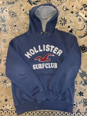 Blå Hollister hoodie med tryck - Blå hoodie från Hollister med stor vit och röd brodyr på bröstet där det står 'Hollister Surfclub'. Hoodien har huva med knappar och mjukt grått foder på insidan. Klassisk känguruficka framtill och ribbade muddar vid ärmar och nederkant. Perfekt för chill dagar.