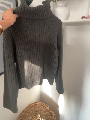 Mörkgrå ribbstickad polotröja - Mysig mörkgrå ribbstickad polotröja med hög krage och långa ärmar. Tröjan har en avslappnad passform och är perfekt för kyliga dagar. Materialet känns mjukt och värmande, och den ribbade strukturen ger en snygg look.