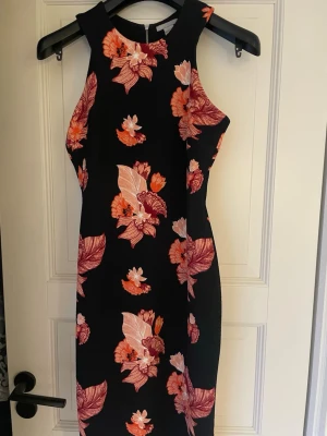 Svart blommig klänning  - Snygg svart kortklänning med färgstarkt blommönster i rosa, orange och rött. Klänningen har ärmlös design, markerade axlar och dragkedja i ryggen. Perfekt för dig som vill sticka ut med en feminin och trendig look. 