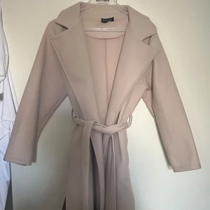 Beige kappa från Missa - Stilren lång beige kappa från Missay med bred krage och knytskärp i midjan. Kappan har en rak passform och är tillverkad i ett mjukt tyg som ger en elegant look. Perfekt för våren och hösten! Använd 1 gång, inga defekter.