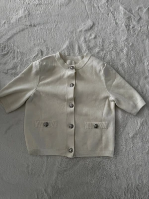 Krämvit kofta med kort ärm - Stilren krämvit kofta med korta ärmar och rund hals. Koftan har dekorativa silverknappar framtill och två små fickor med knappdetalj. Ribbstickade muddar vid ärmslut, nederkant och hals. Perfekt för en clean och enkel look.