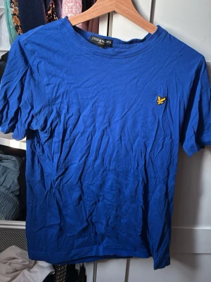 Blå t-shirt från Lyle & Scott - Klassisk blå t-shirt från Lyle & Scott med rund hals och korta ärmar. T-shirten har den ikoniska gula logon broderad på bröstet och är tillverkad i mjuk bomull. Perfekt basplagg för en enkel och stilren look.