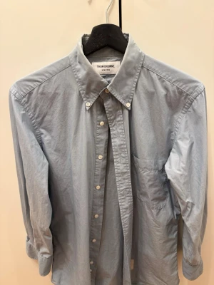 Ljusblå skjorta från Thom Browne - Klassisk ljusblå skjorta från Thom Browne med button-down krage och bröstficka. Skjortan har vita knappar framtill och på kragen, samt långa ärmar. Perfekt för en clean och stilren look.