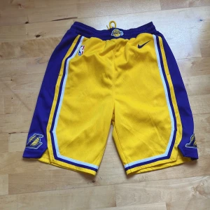 Lakers basketshorts från Nike XS - Gula och lila basketshorts från Nike med Los Angeles Lakers-logga och NBA-märke. Shortsen har elastisk midja, vita och blå ränder längs sidorna och är tillverkade i ett lätt polyestermaterial. Perfekta för basket eller sportiga outfits.