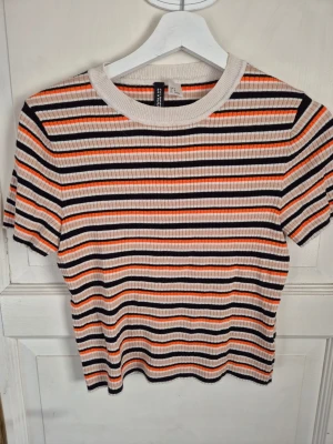 Randig ribbad topp från H&M - Säljer en ribbad topp från H&M med korta ärmar och rund hals. Toppen har ett färgglatt randigt mönster i orange, svart, beige och vitt. Perfekt för dig som gillar en retroinspirerad stil med lite edge.