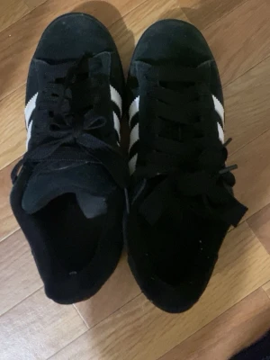 Svarta Adidas sneakers med vita ränder - Klassiska svarta Adidas sneakers med tre vita ränder på sidan. Skorna har låg profil och är gjorda i mockaliknande material med svart sula och svarta skosnören. Perfekta för en stilren och sportig look.