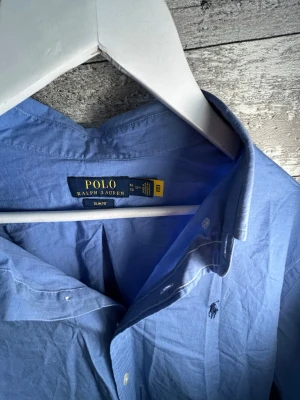 Blå skjorta från Polo Ralph Lauren - Klassisk blå skjorta från Polo Ralph Lauren med button-down krage och broderad logga på bröstet. Skjortan har långa ärmar, knäppning framtill och är tillverkad i bomull. Perfekt för en stilren look.