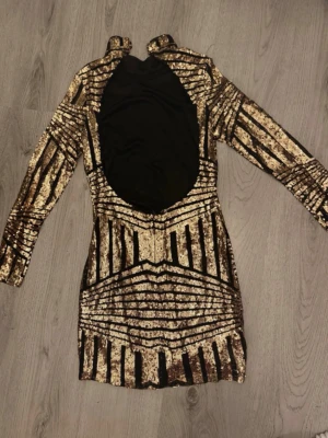 Glittrig guldklänning med lång ärm - Snygg kort klänning i guld och svart med glittrigt mönster och lång ärm. Hög hals och öppen rygg ger en extra cool vibe. Perfekt för dig som vill sticka ut! Materialet är troligtvis polyester med paljettdetaljer.