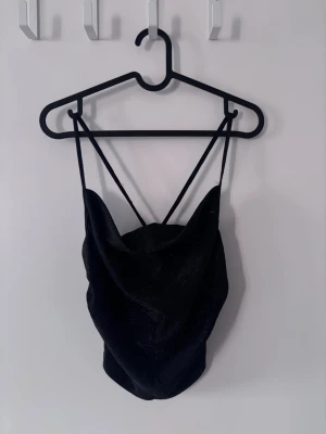 Svart glittrig linnetopp med tunna band - Snygg svart linnetopp med tunna axelband och glittrigt tyg. Toppen har en draperad framsida som ger en cool och trendig look. Passar till många olika stilar och är lätt att matcha.