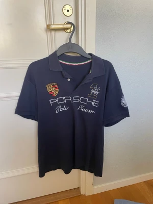 Mörkblå Porsche pikétröja - Snygg mörkblå pikétröja från Porsche med broderade detaljer och logga på bröstet samt texten 'Porsche Polo Team'. Klassisk krage och korta ärmar. Perfekt för dig som gillar sportig stil och exklusiva märken.