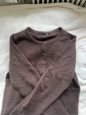 Brun kofta från lager 157 med knappar - Mysig brun kofta från lager 157 med rund halsringning och tre knappar framtill. Tillverkad i ett mjukt, luddigt material som ger en avslappnad look. Perfekt att slänga över en t-shirt eller linne för extra värme.