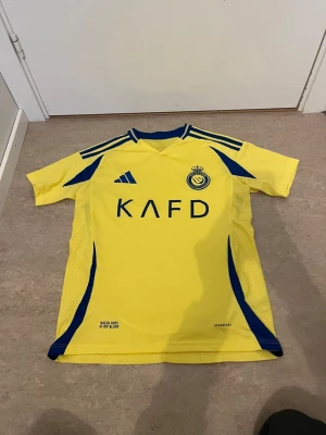 Gul Al Nassr matchtröja Adidas - Säljer en gul Al Nassr matchtröja från Adidas med blå detaljer och klubbmärke på bröstet. Tröjan har korta ärmar, blå ränder på axlarna och tryckt sponsorlogga framtill. Tillverkad i lätt och ventilerande material, perfekt för fotbollsträning.