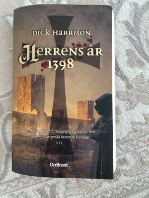 Herrens år 1398 av Dick Harrison - En historisk roman av Dick Harrison med titeln 'Herrens år 1398'. Boken har ett omslag i bruna och beige toner med motiv av borg, kors och en munk. Tryckt på vanligt vitt papper och utgiven av Ordfront. Perfekt för dig som gillar medeltida berättelser.