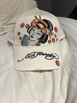 Vit Ed Hardy keps med geisha-brodyr - Säljer en vit keps från Ed Hardy med broderad geisha och blommor i orange, rött och gult framtill. Kepsen har mesh-bakstycke och justerbar passform. Märkets logga är broderad på skärmen och en liten blomma finns även bak. Perfekt statement piece för din streetwear-look.