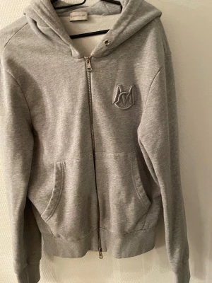 Grå zip hoodie från Moncler - Snygg grå hoodie från Moncler med dragkedja framtill och broderad logga på bröstet. Hoodien har huva, ribbade muddar och känguruficka. Tillverkad i mjuk bomullsblandning som känns riktigt skön mot huden. Perfekt för en avslappnad och trendig look. Passar även S  