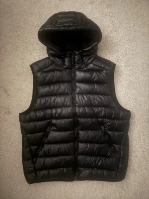 Svart dunväst med huva - Snygg svart dunväst med huva och dragkedja framtill. Västen har två snedställda fickor med dragkedja och quiltad design med horisontella sömmar. Perfekt för lager-på-lager och ger en sportig look.