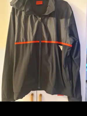 Svart vindjacka från Hugo med röd detalj - Snygg svart vindjacka från Hugo med dragkedja, huva och röd linje som går runt bröstet och ryggen. Jackan har en sportig look och är tillverkad i lätt, vindtätt material. Perfekt för dig som gillar stilrena och moderna plagg.