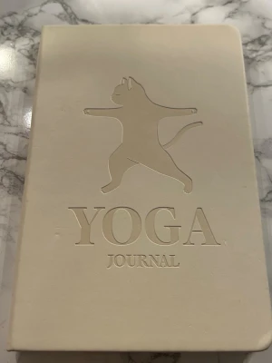 Beige Yoga Journal med kattmotiv - Säljer en beige Yoga Journal med ett präglat kattmotiv på framsidan. Omslaget är mjukt och boken har sidor för veckoplanering, affirmationer och anteckningar. Perfekt för dig som vill hålla koll på din yoga och reflektioner.