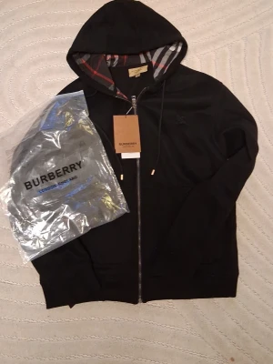 Svart hoodie från Burberry - Snygg svart hoodie från Burberry med dragkedja och klassiskt rutigt foder i huvan. Märkets logga är diskret broderad på bröstet. Tillverkad i mjukt material och har ribbade muddar. Perfekt för dig som vill ha en stilren och exklusiv look. Har alla tags.