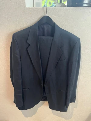 Svart kavaj från Pierre Cardin - Säljer en stilren mörkblå kostym från Pierre Cardin i linne. Klassisk modell som passar perfekt både till mer uppklädda tillfällen och smart casual outfits. Tidlös design med enkel knäppning och rena linjer. Fungerar bra både till kostymbyxor, chinos eller jeans. Använd ett fåtal gånger så fortfarande i fint skick. Märke: Pierre Cardin Färg: Mörkblå Storlek: ca L / 52 Skriv gärna om du har frågor!