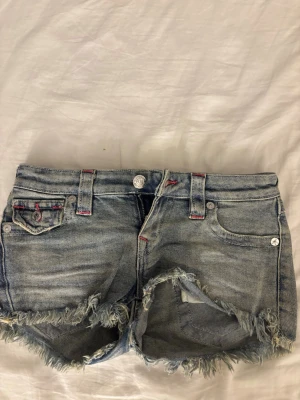 True Religion jeansshorts med fransar - Säljer ett par ljusblå jeansshorts från True Religion med fransig kant. Shortsen har röda sömmar och är low waist. Säljer för jag inte använder dom så dom bara ligger i garderoben. Nypris 1200 ungefär, priset kan diskuteras.