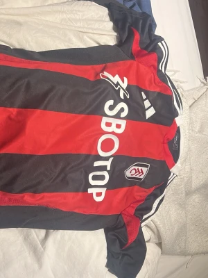 Fulham FC matchtröja Adidas röd/svart - Snygg Fulham FC matchtröja från Adidas i rött och svart med vita detaljer. Tröjan har klubbmärke på bröstet, SBOTOP-logga och klassiska Adidas-ränder på axlarna. Perfekt för fotbollsträning eller att bära till vardags. Köpt i london