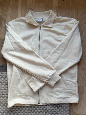 Ziptröja från Zara - Stilren beige ziptröja från Zara med ribbad krage och långa ärmar. Tröjan har dragkedja framtill och är tillverkad i mjukt bomullsmaterial. Perfekt för en clean och avslappnad look.