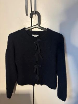 Mörkblå kofta med rosettknäppning - Snygg mörkblå kofta med långa ärmar och rosettknäppning framtill. Koftan har en kortare passform och är gjord i ett mjukt, fluffigt material som känns riktigt cozy. Perfekt för att lyfta din outfit med en unik detalj.