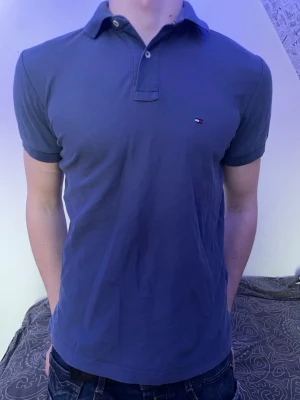 Blå pikétröja från Tommy Hilfiger - Klassisk blå pikétröja från Tommy Hilfiger med liten logga på bröstet. Tröjan har krage, korta ärmar och knappar vid halsen. Perfekt till jeans eller shorts för en clean och stilren look.